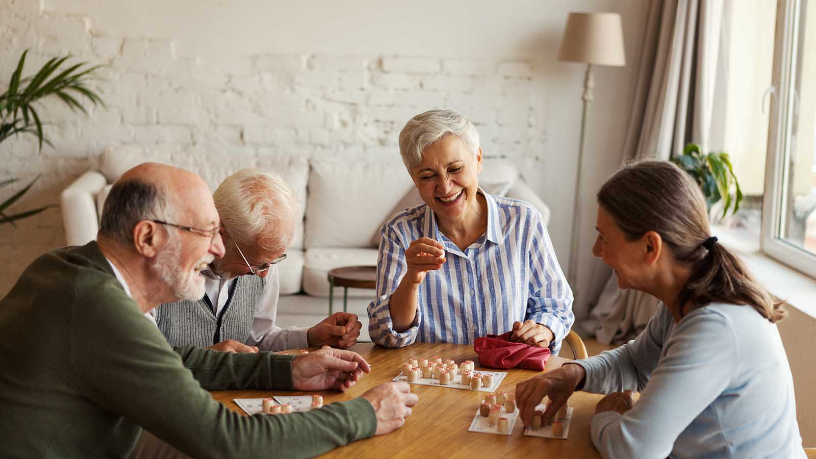 Group of four cheerful senior people, two men and two women, having fun sitting at table and playing bingo game in nursing home