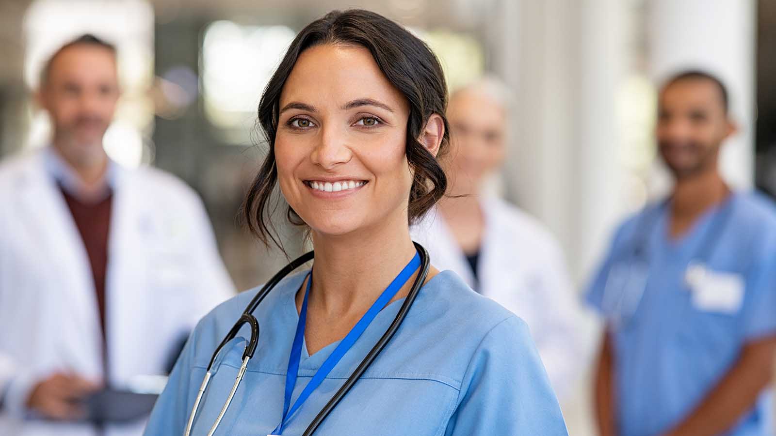 Portrait of smiling young nurse in uniform smiling with healthcare team in background.